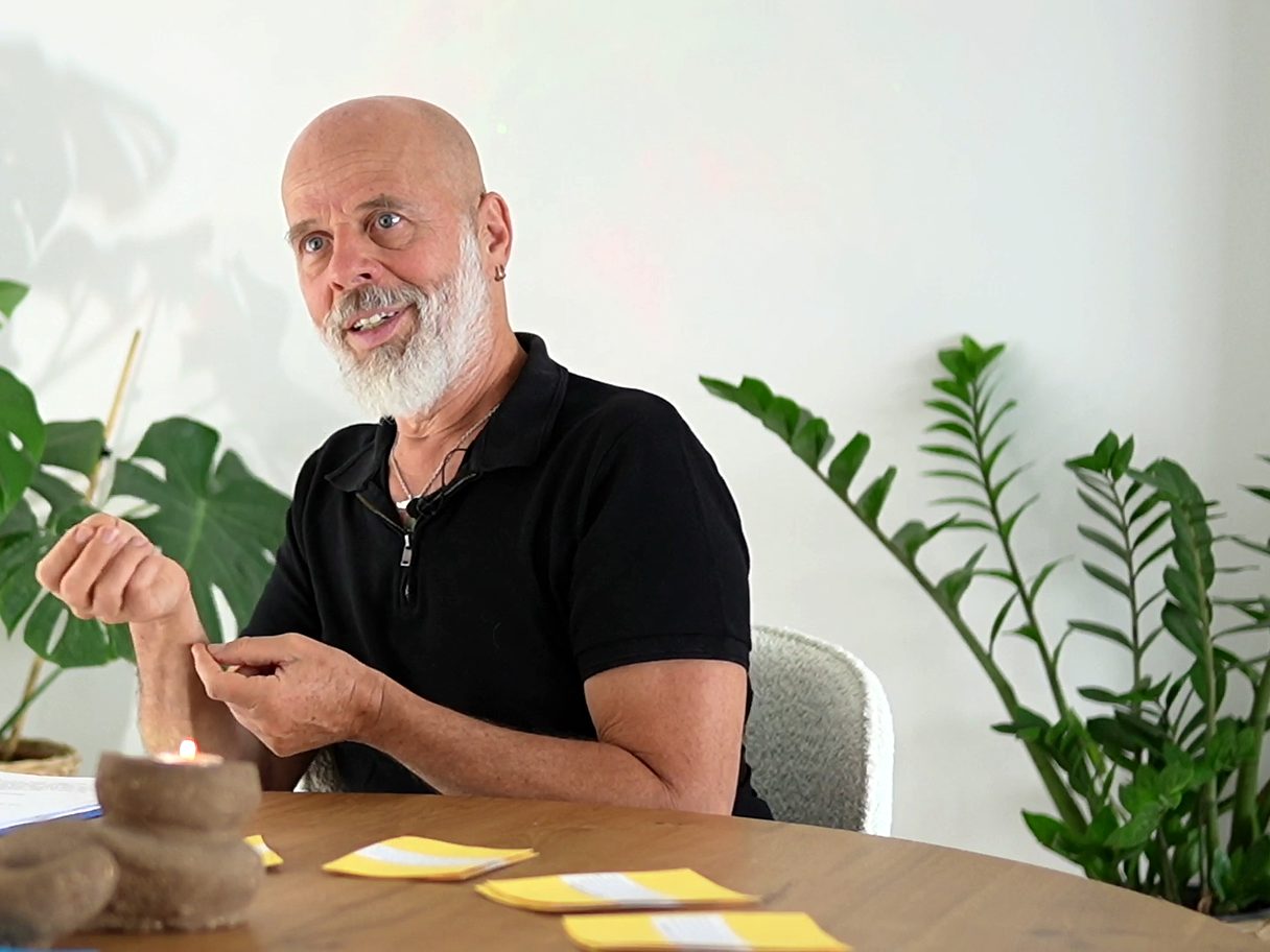 זהר וילסון מדבר ואוחז בידו, יושב ליד שולחן עץ ומאחוריו צמחייה ירוקה